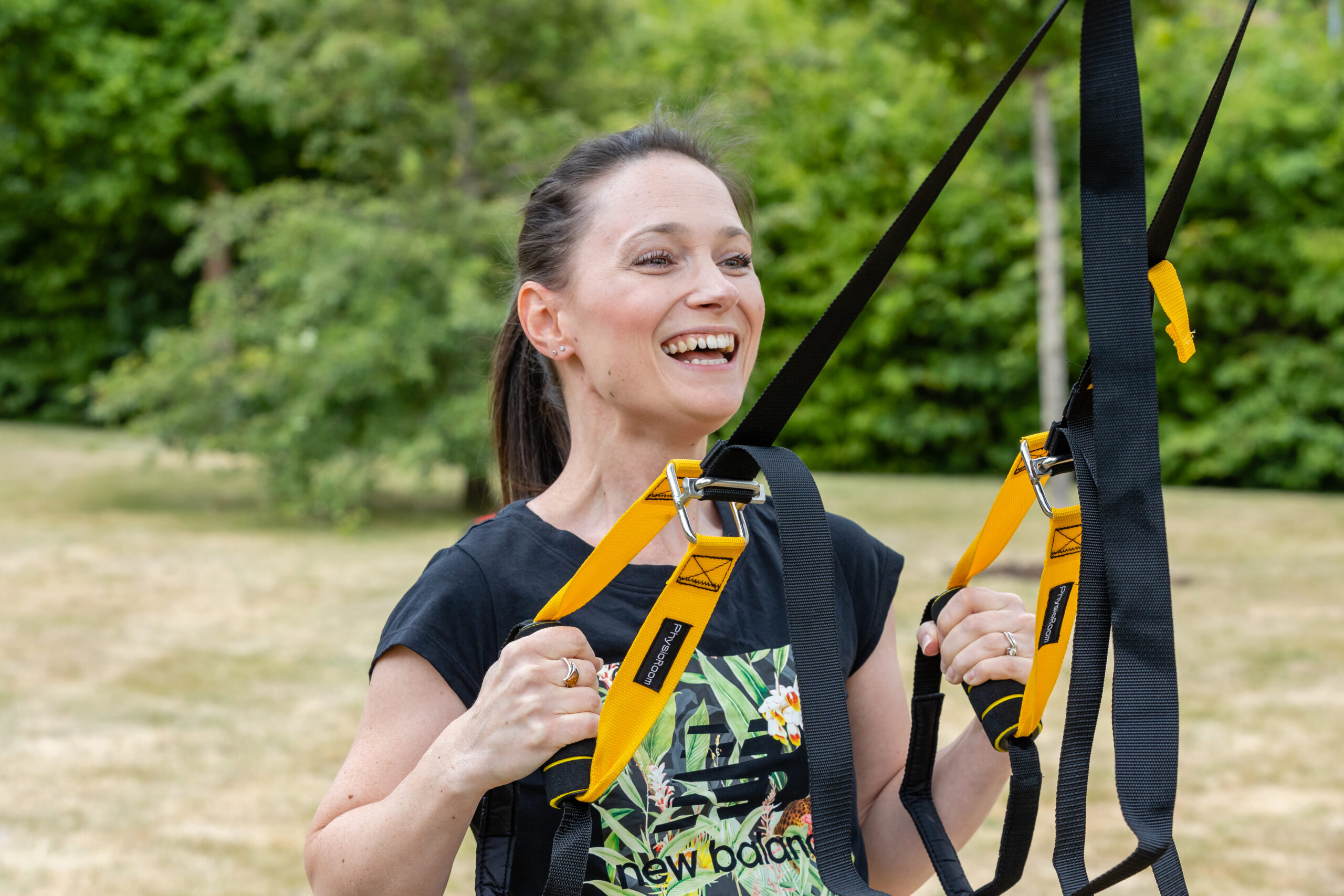 A woman smiling and holding onto yellow and black suspension training straps outdoors, with green trees in the background.