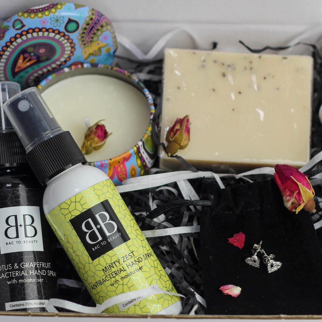 A gift box containing a bar of soap, a candle, a bottle of citrus hand spray, a bottle of mint spray, a pair of earrings in a velvet pouch, and dried rosebuds on black shredded paper.