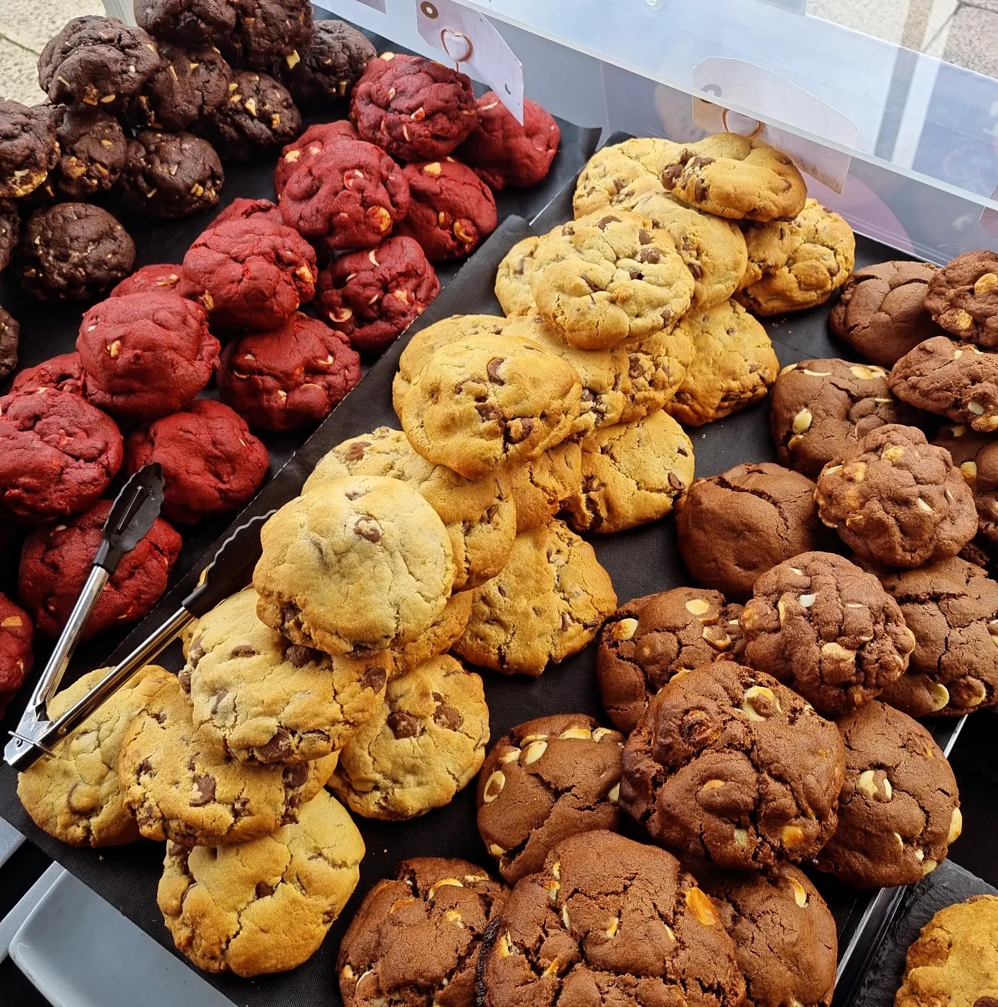 Rows of assorted biscuits including chocolate chip, double chocolate, red velvet, and peanut butter, displayed in a tray at a bakery or market.