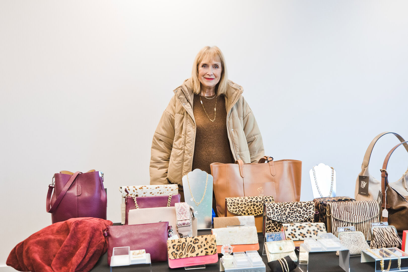 A woman stands behind a table displaying various handbags, wallets, and jewellery against a plain white background.