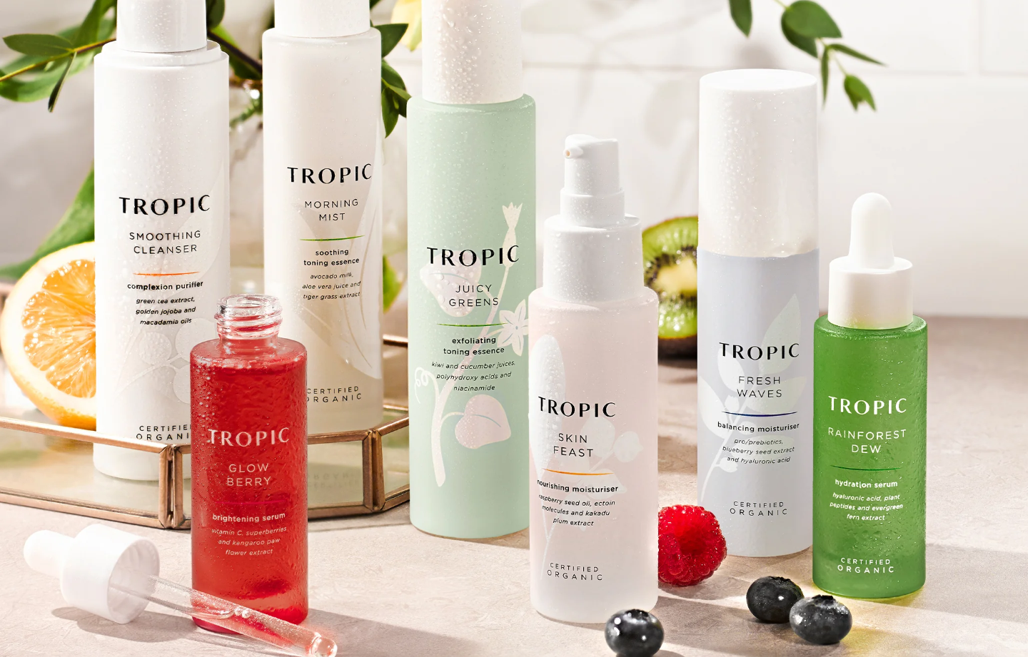 A collection of Tropic skincare products, including cleansers, mists, serums, and moisturisers, arranged on a surface with fruits like orange, kiwi, and berries nearby.