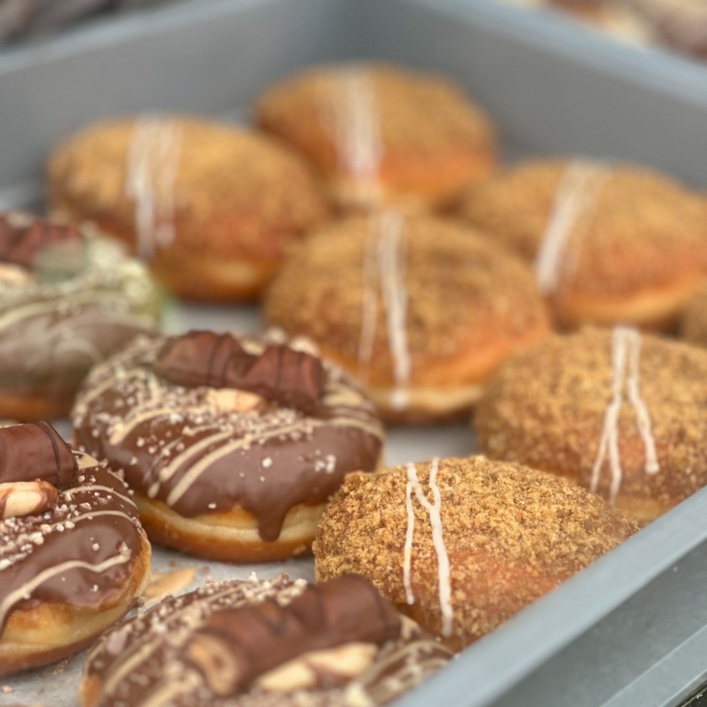 A tray of assorted doughnuts, some topped with chocolate and sweet pieces, others with a crumb coating and white icing drizzle.